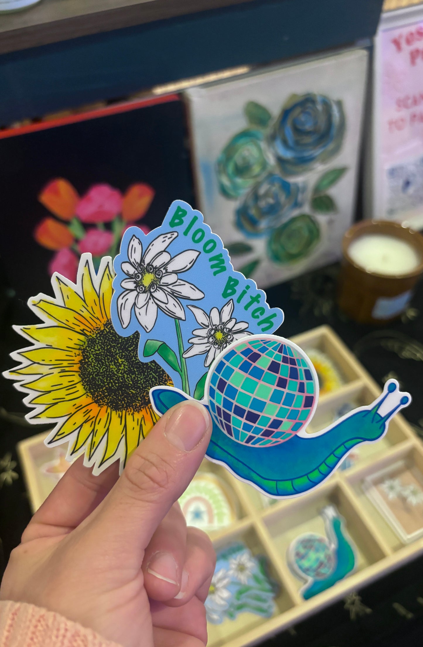 Hand holding a colorful sticker with a sunflower, flower, and snail design in front of a decorative background.